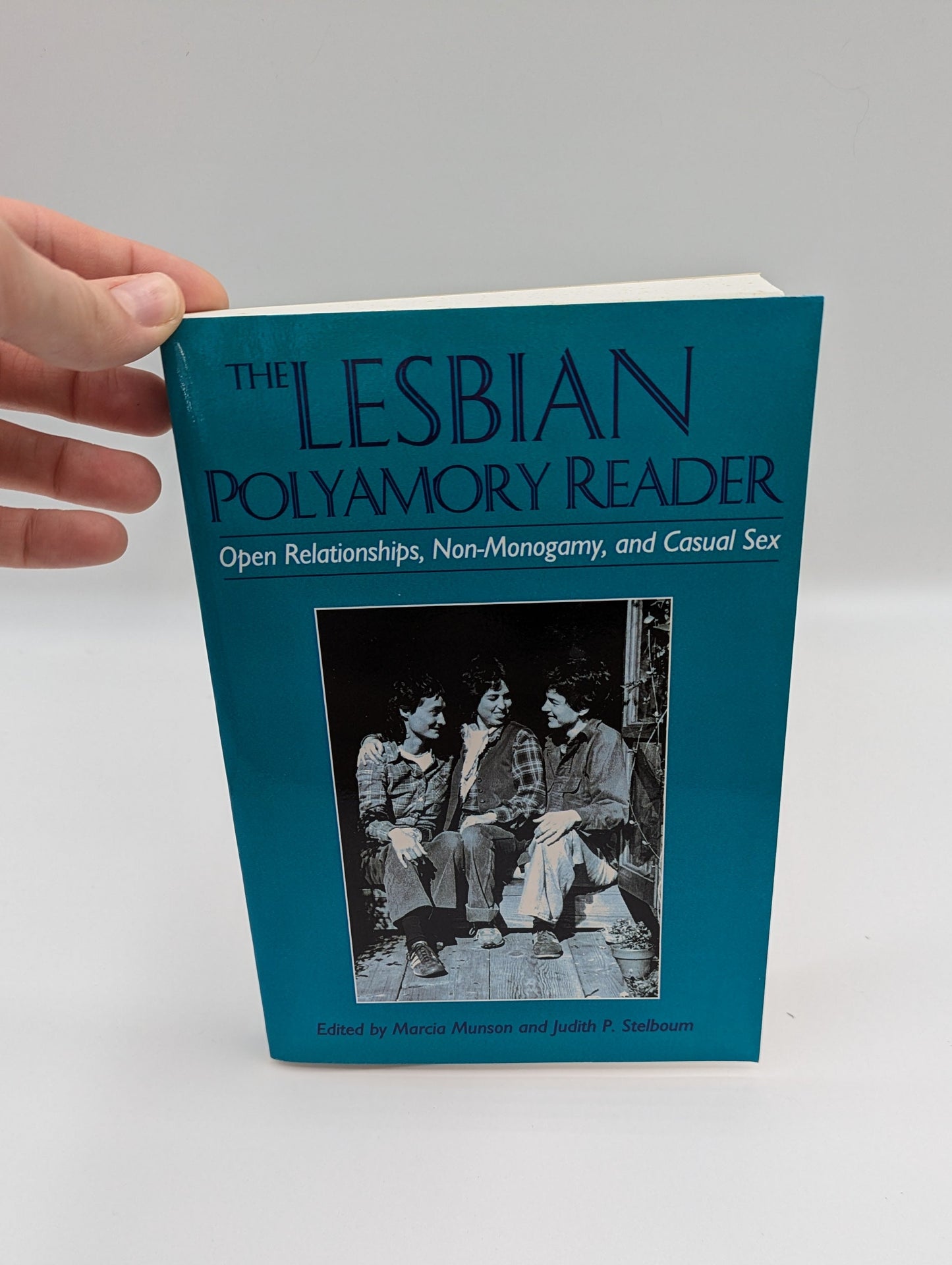 Book titled 'The Lesbian Polyamory Reader' held by a hand on a white background
